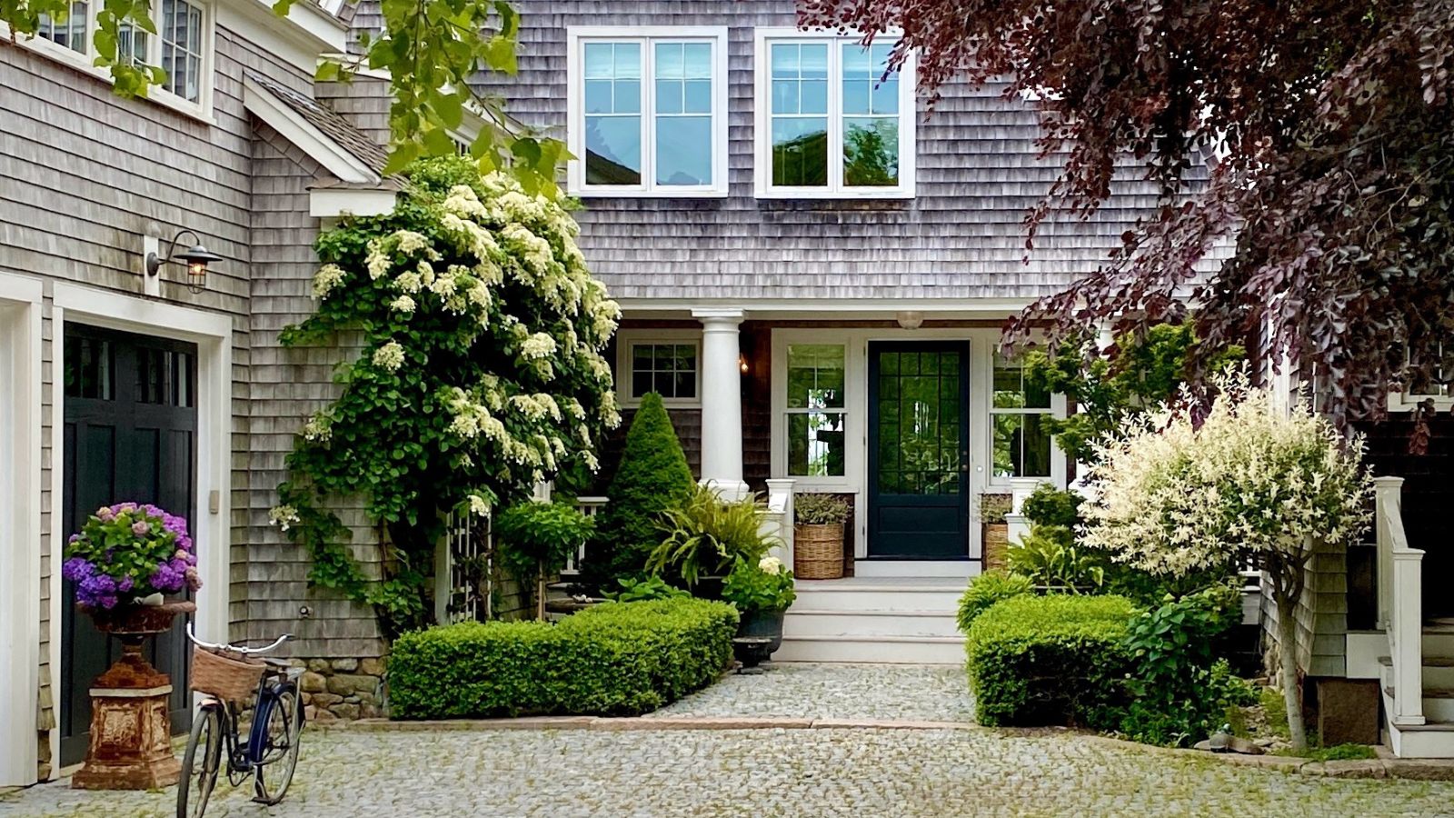 Shingle style house with front porch with planters, hedges, and two garages with a bicycle outside
