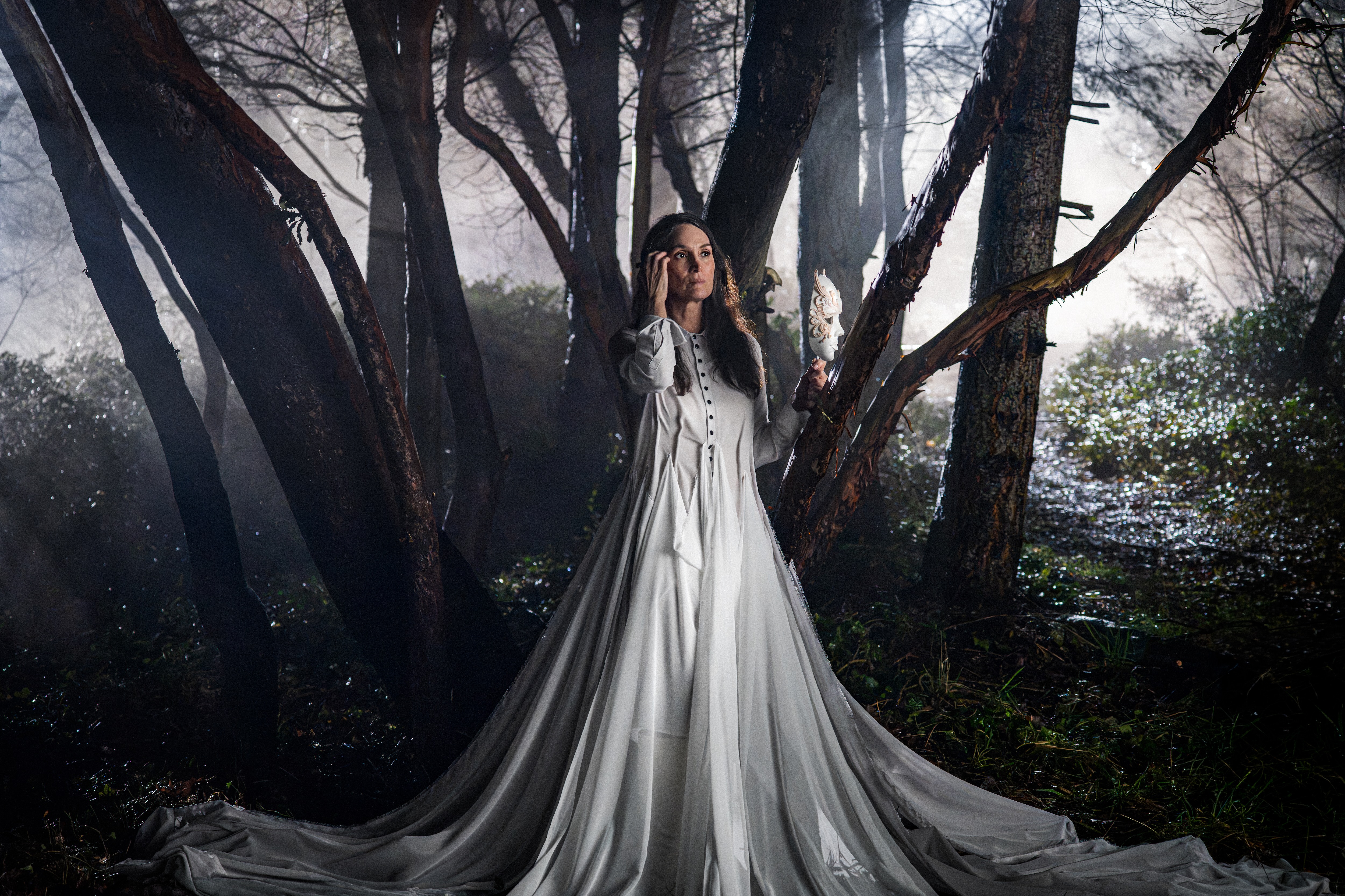 Carrie Anne Moss wears a white gown while standing in a mythical forest with a white light shining behind the trees, in the Crave TV show 'Yaga.'
