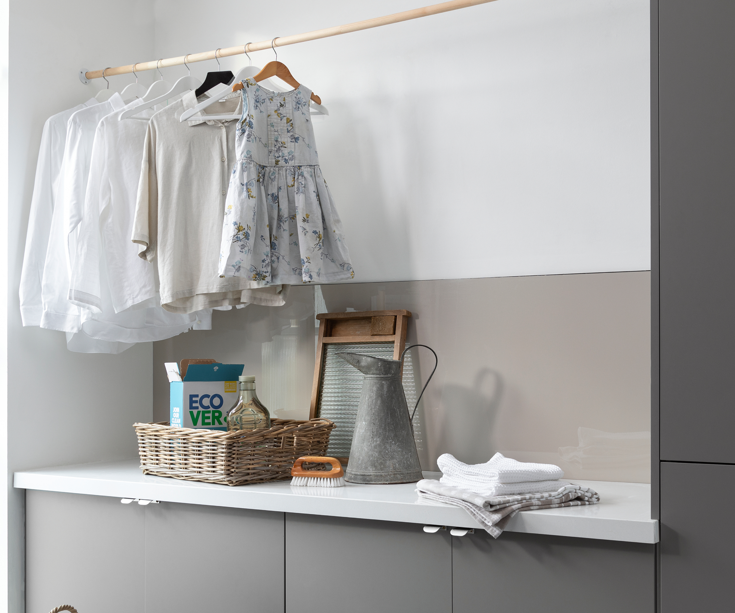 laundry room with white walls, metal splashback. hanging rail wth clothes on and laundry materials on white worktop with grey cabinets below