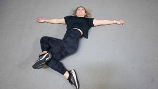 Woman lies on her back with her knees bent and legs on the floor to her right-hand side, her right foot resting on her left knee. She wears a black T-shirt, black leggings and black trainers.