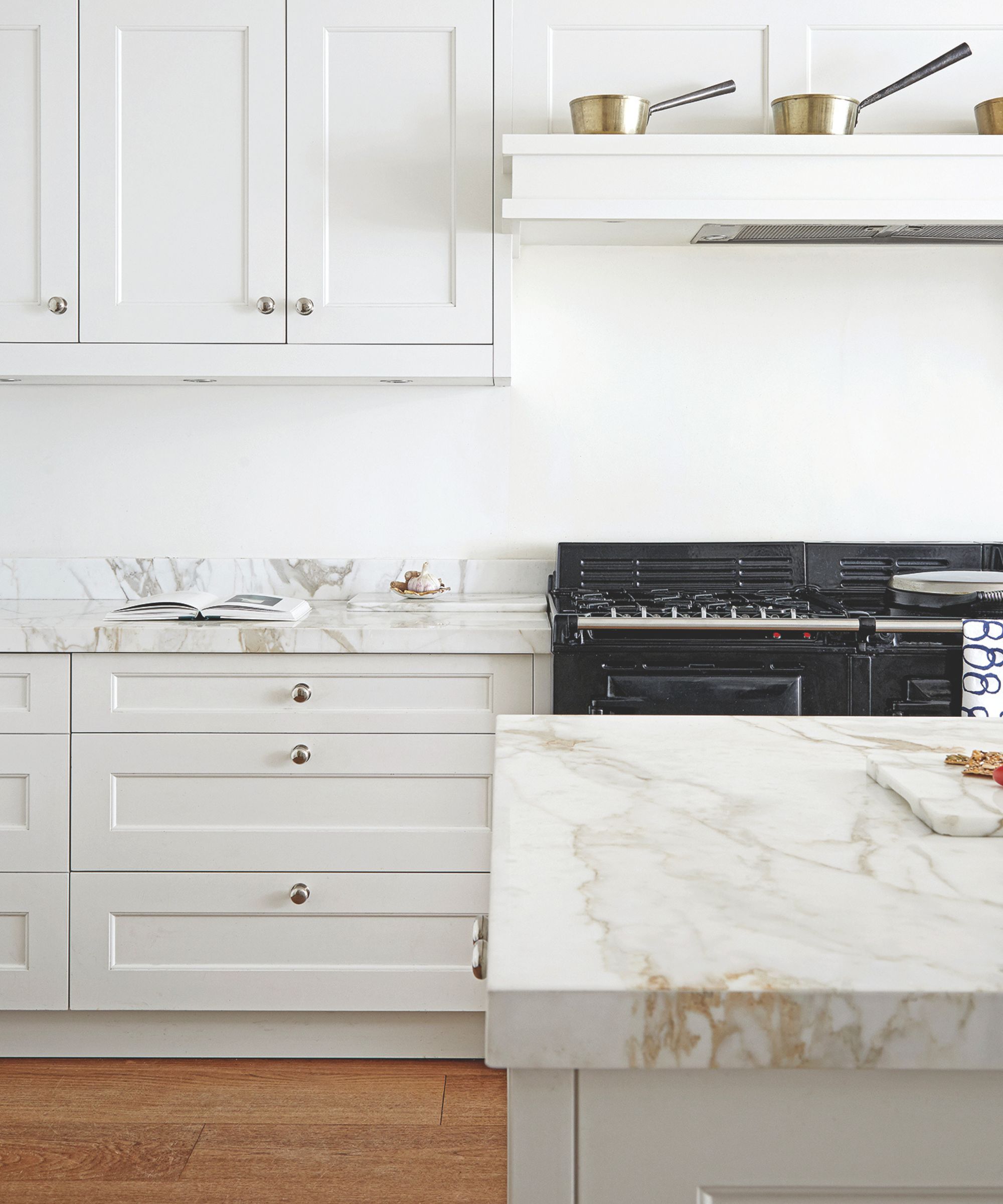 A white kitchen with white cabinetry, wooden floors, and white marble countertops. There&#039;s a large black AGA stove, and shelving above with copper pans.
