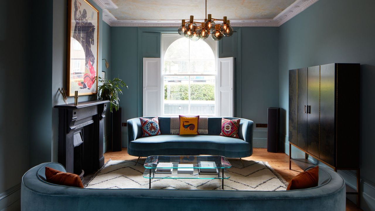 blue living room with two curved blue sofas facing each other, a glass coffee table, an original fireplace with art hanging over it, a cabinet on the opposite wall, and a modern pendant light