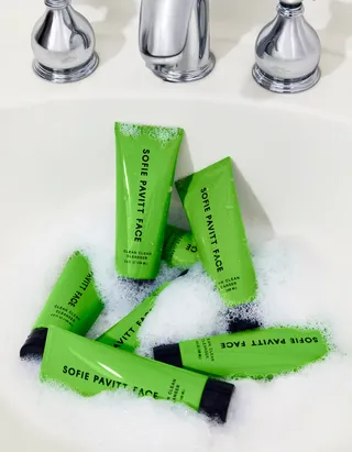 Multiple tubes of face cleanser in a bright green color lay in a white and chrome sink filled with foam.