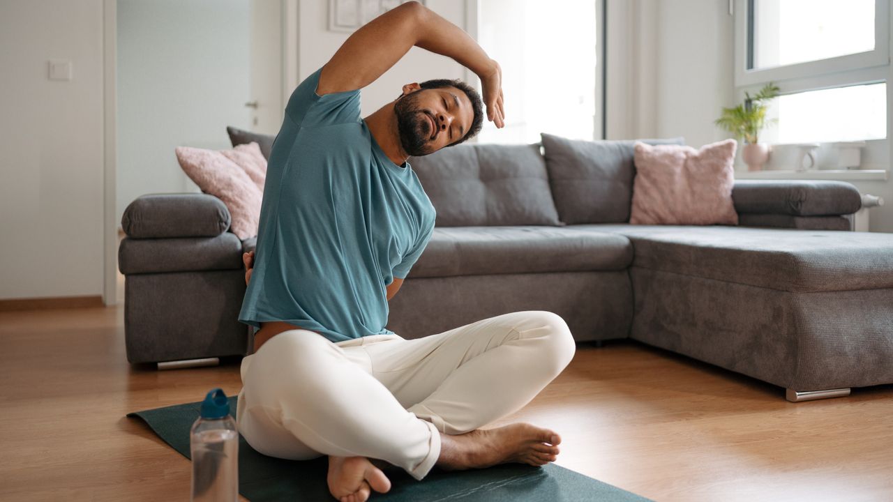 A man sits on a yoga mat performing stretches in a living room. He is cross-legged, with his left hand behind him for balance and his right arm bent and reaching over his head. His eyes are closed and he wears a t-shirts and soft trousers. Behind him is an L-shaped couch with lots of cushions and a small plant.