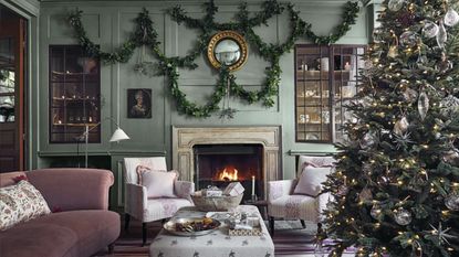 Living room with green walls, green mantelpiece and lit fire with armchairs either side, central ottoman beside decorated Christmas tree.