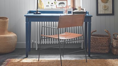 Blue desk with leather chair in front of white panelled wall with jute carpet, white radiator, lemon artwork, and mismatched storage baskets