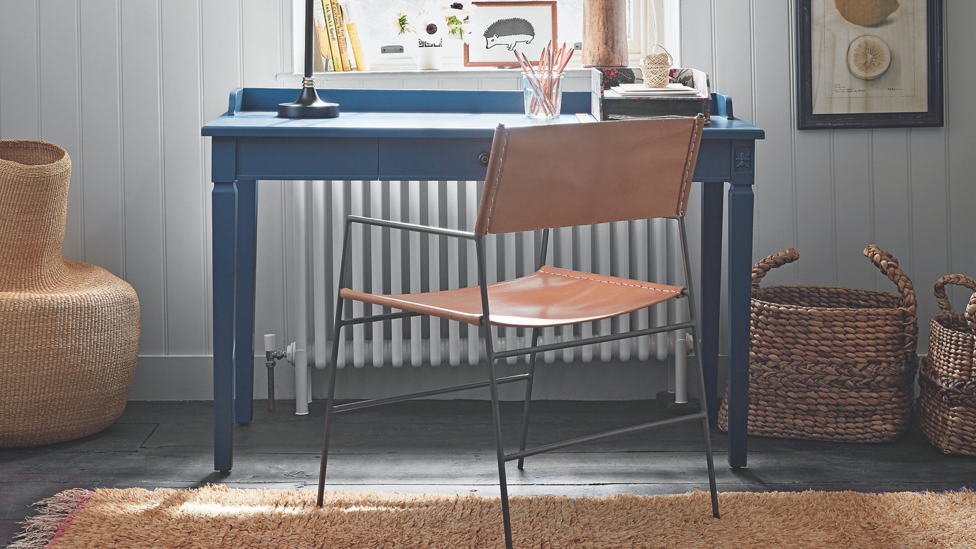 Blue desk with leather chair in front of white panelled wall with jute carpet, white radiator, lemon artwork, and mismatched storage baskets