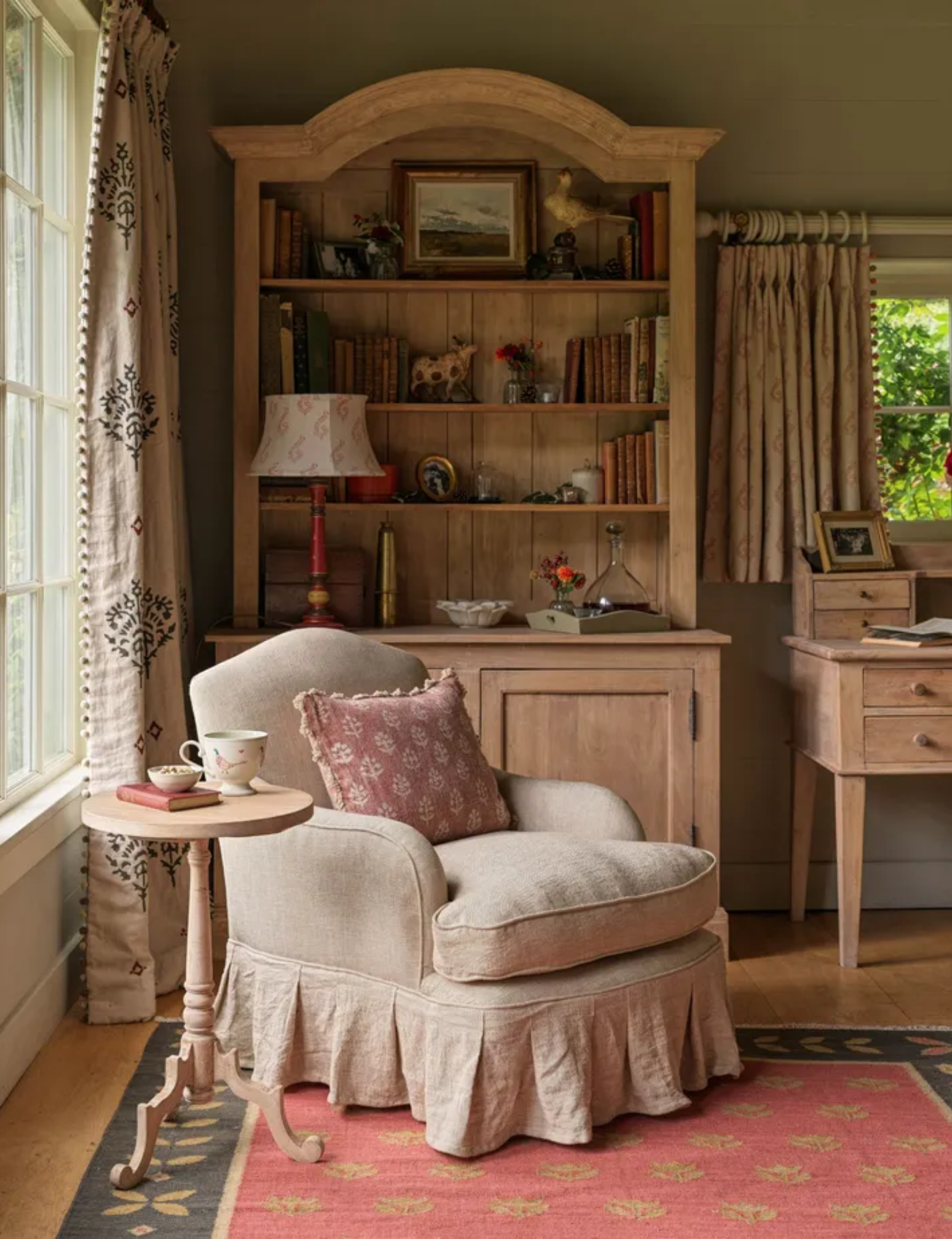 A relaxing reading nook featuring a beige skirted armchair with a pink cushion next to a tall wooden bookcase.