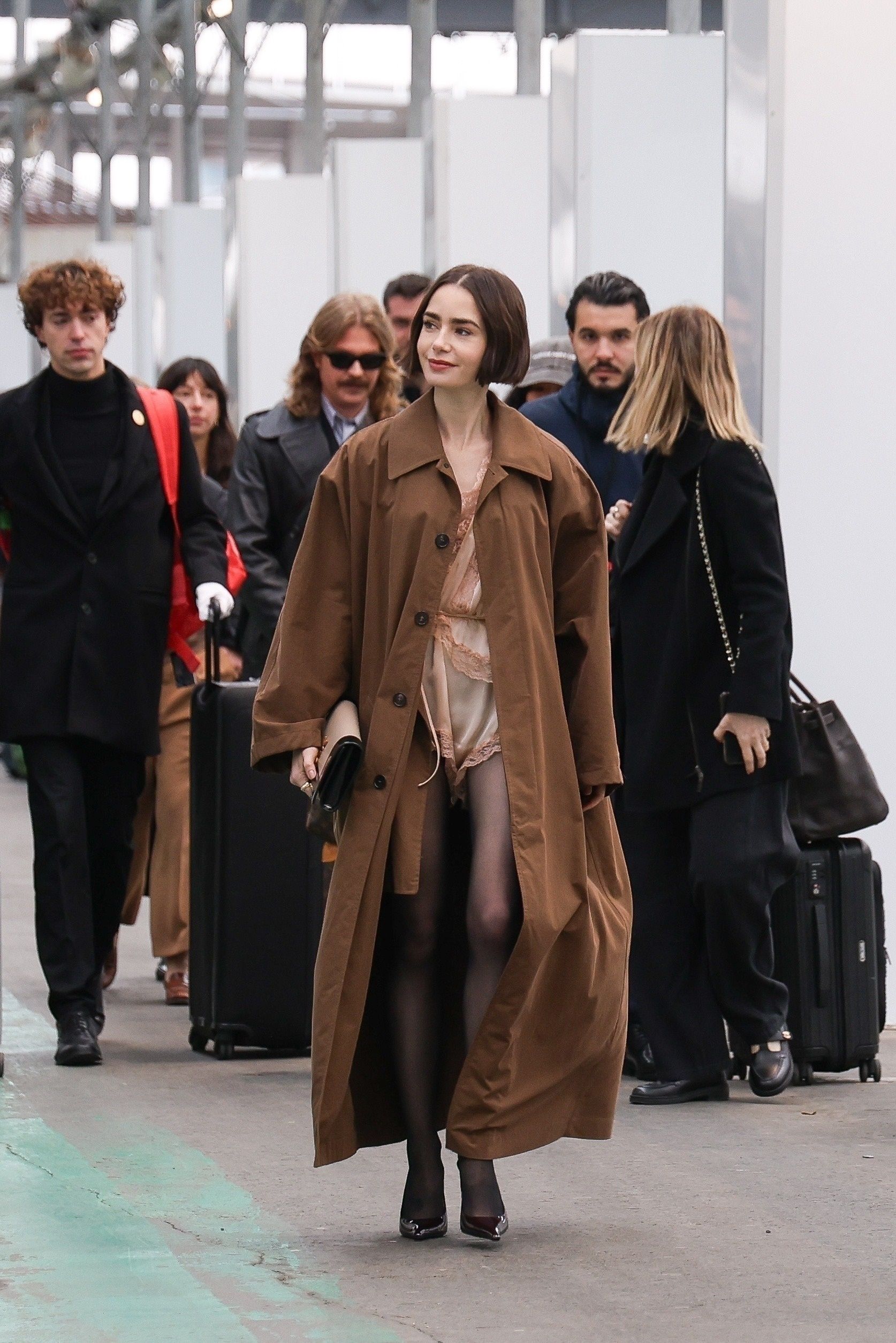 Lily Collins arrives at Gare d’Austerlitz in Paris wearing a lingerie romper from YSL with a trench coat, tights, and heels, for Emily in Paris.