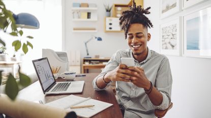 A young man smiles while looking at his phone while sitting at his desk in front of a laptop that shows graphs.