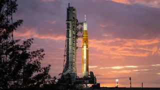 rocket on a launch pad in front of a red sky and with trees in front