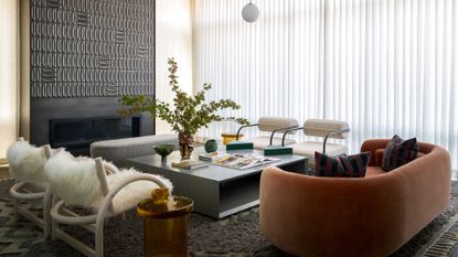 A white living room with sheer curtains, a black tiled accent wall with an in-built fireplace, a gray coffee table, a round amber couch, a pair of metal tube chairs with white round cushioning
