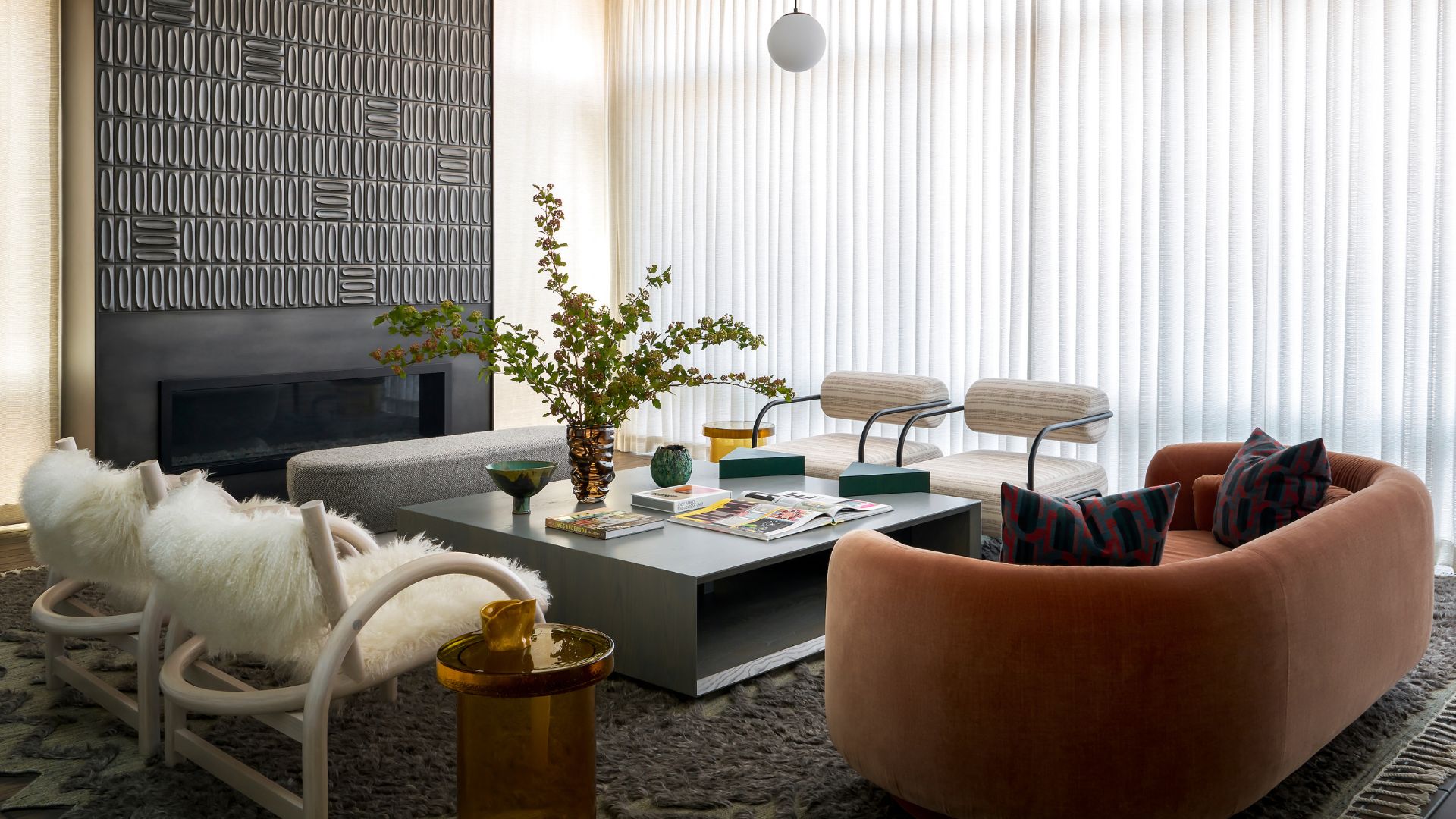 A white living room with sheer curtains, a black tiled accent wall with an in-built fireplace, a gray coffee table, a round amber couch, a pair of metal tube chairs with white round cushioning