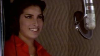 Amy Winehouse smiling in a recording booth with a microphone beside her.