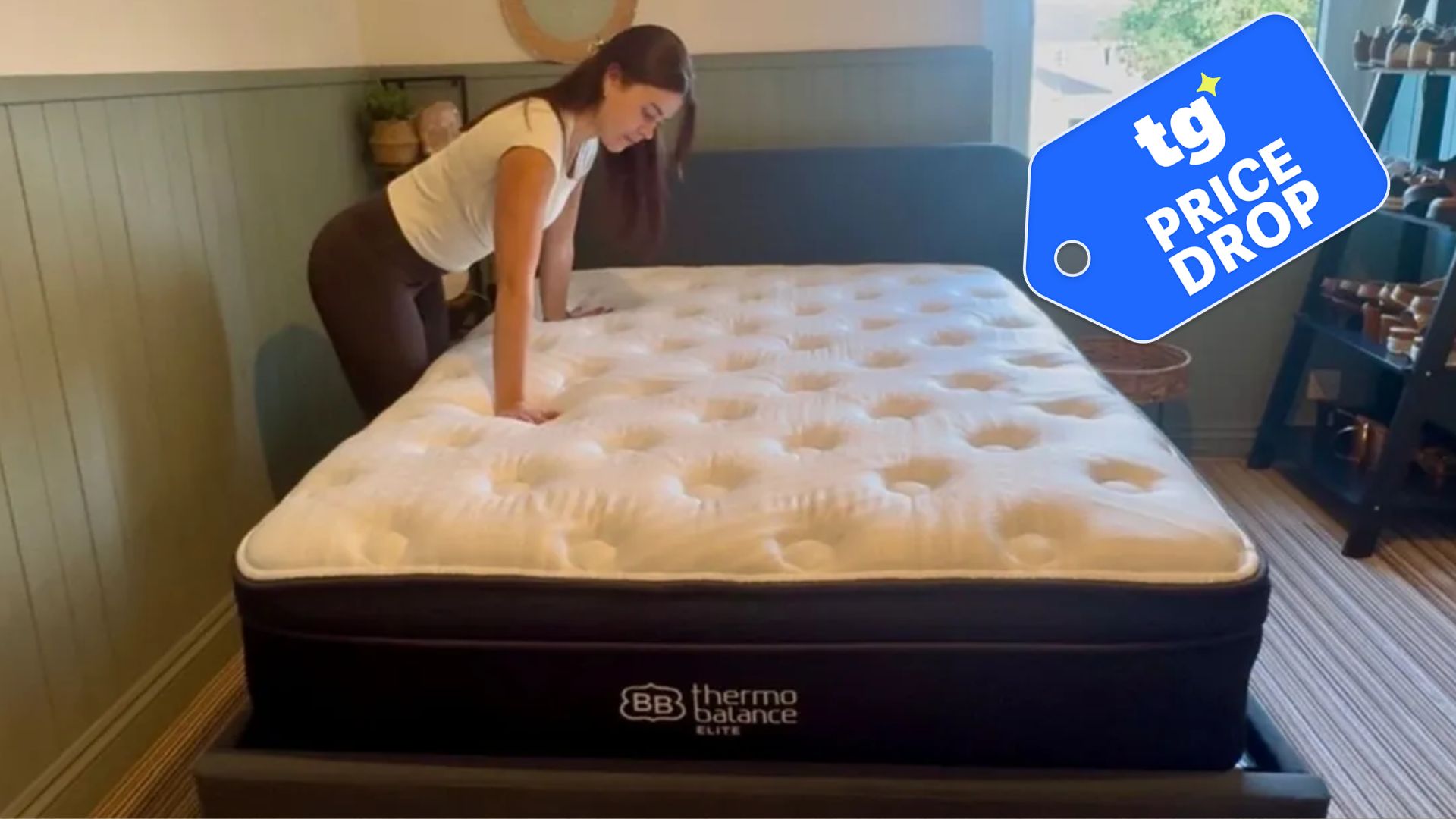 A dark-haired Tom's Guide mattress tester stands next to a blue mattress with a white cover, placing her hands on the surface. Overlaid on top is a Tom's Guide 'Price Drop' label
