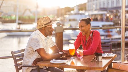 A happy older couple hold hands at an outdoor table at a casual restaurant. There are boats in the background.