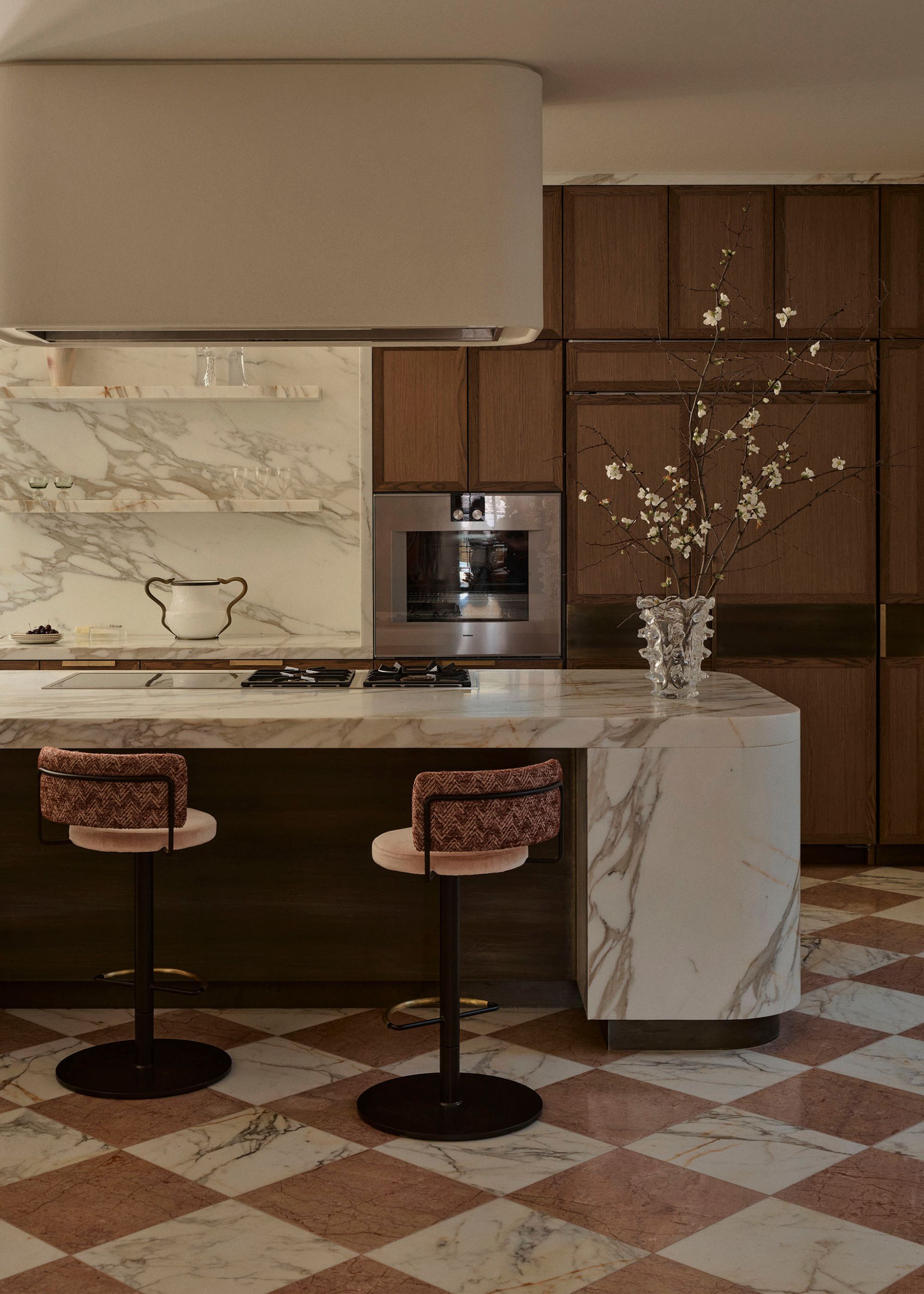 A modern kitchen with checkerboard marble flooring, a stone island, patterned swivel stools, a textured glass vase with stems, and brown cabinetry