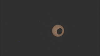 A yellowish sun against a black background is partially obscured by a Martian moon. 