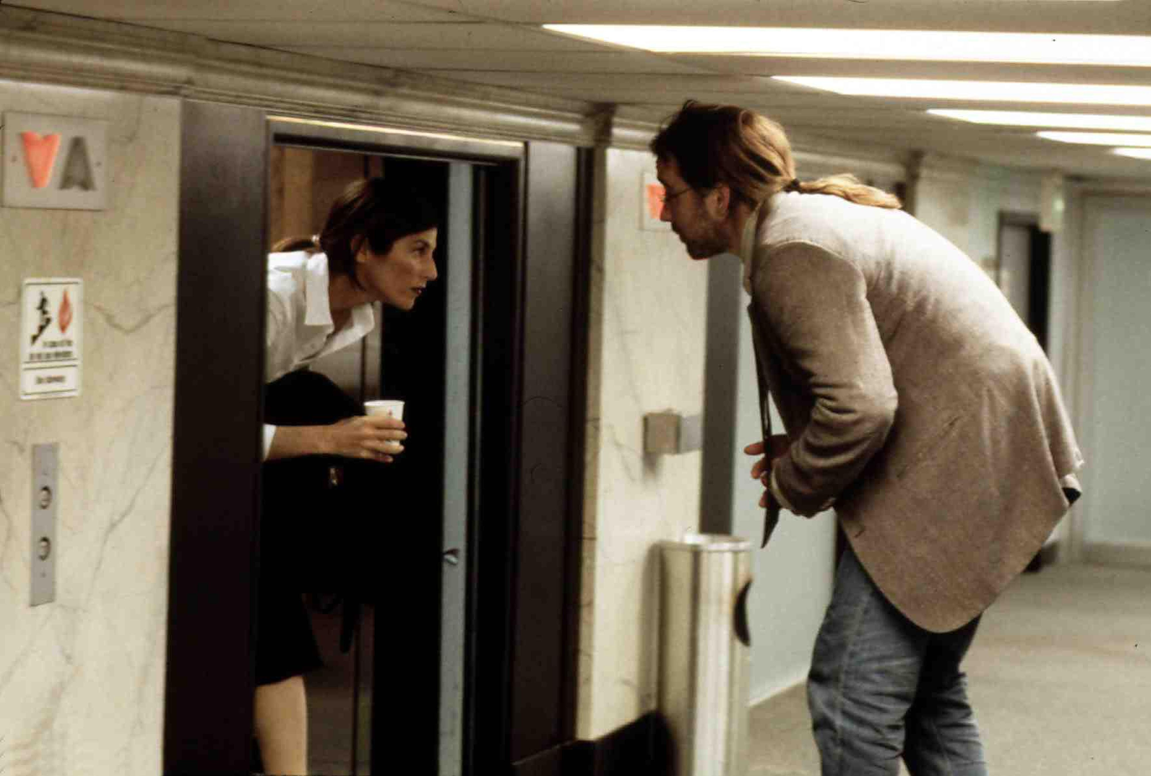 (L to R) Catherine Keener as Maxine and John Cusack as Craig outside of an elevator in a too-short hallway that forces them to crouch in Being John Malkovich