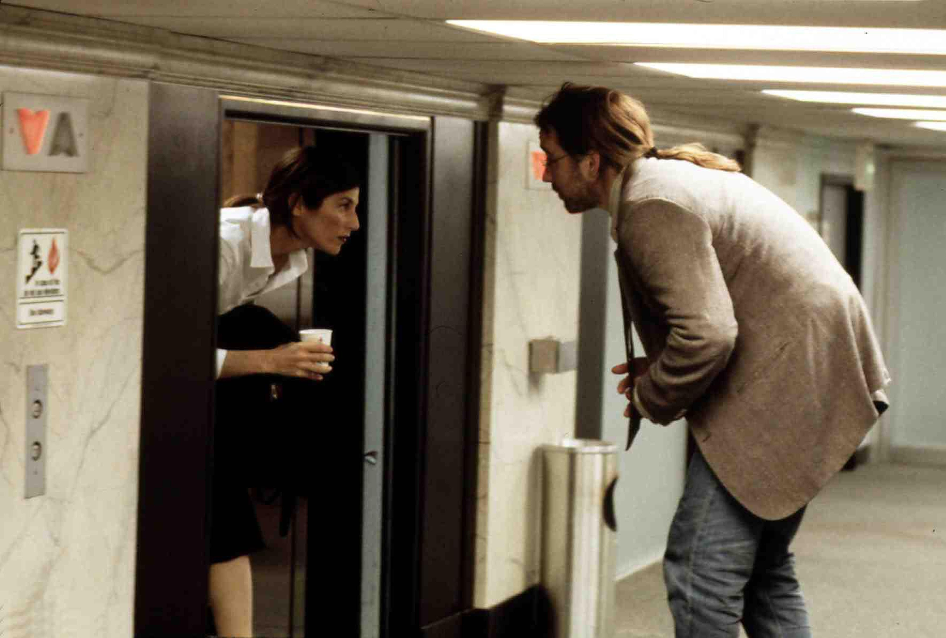 (Von links nach rechts) Catherine Keener als Maxine und John Cusack als Craig vor einem Aufzug in einem zu kurzen Flur, der sie dazu zwingt, sich in „Being John Malkovich“ zu ducken