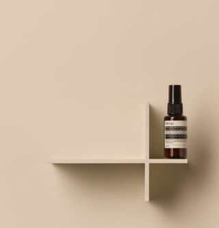 A bottled mist with a black and white label sits on an empty wooden shelf against a cream background.