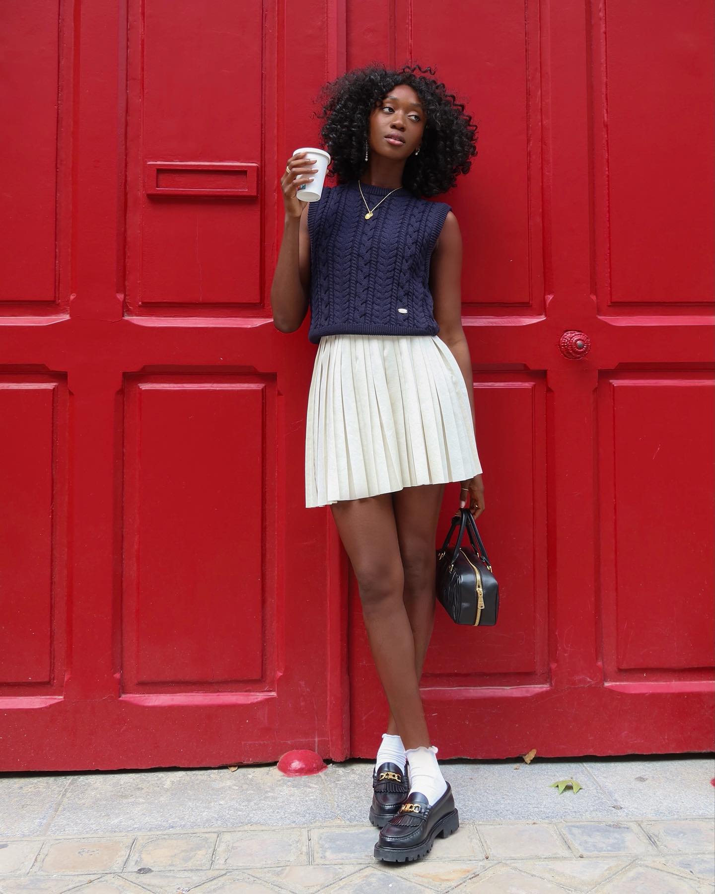 French style influencer posing with a cup of coffee in front of red doors in Paris wearing a sleeveless navy sweater vest, pleated white skirt, ruffle socks, and black chunky loafers