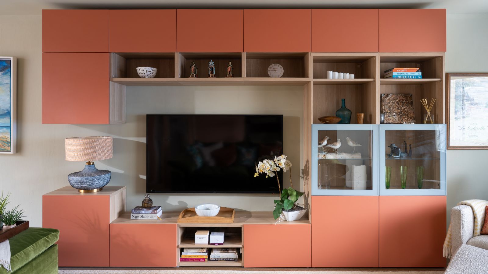 Living room with built-in cubby storage on the wall painted in Naperon by Farrow &amp; Ball, a large TV mounted on the wall at the centre, and a lamp and orchid on either side.