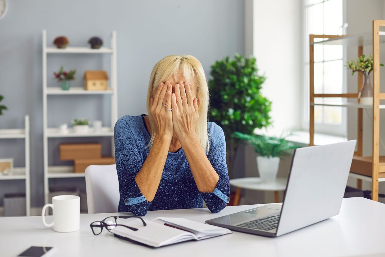 A woman puts her hands over her face while sitting in front of a laptop.