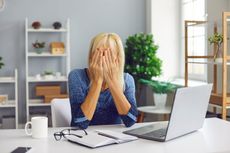 A woman puts her hands over her face while sitting in front of a laptop.