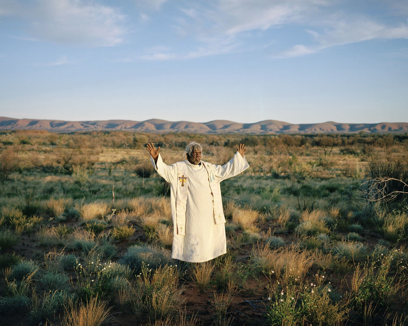 A pastor adorned in white robe with arms outstretched in the Australian outback