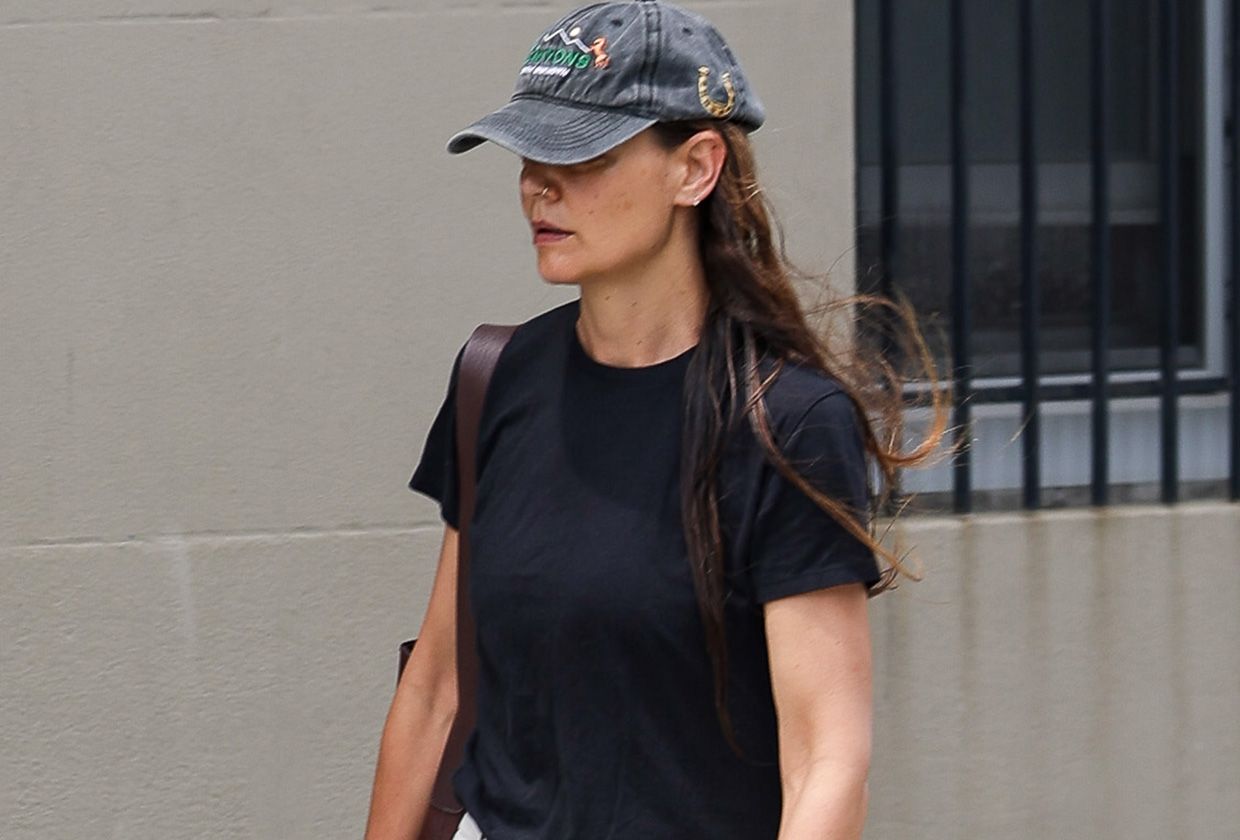 Katie Holmes walking in New York City wearing a baseball cap, black T-shirt, white trousers, and almond-toe black flats.