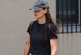 Katie Holmes walking in New York City wearing a baseball cap, black T-shirt, white trousers, and almond-toe black flats.