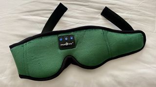 A closeup of the MUSICOZY Headphone Sleep Mask in green against a white sheet with the straps showing at the back