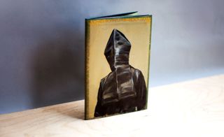 A hardcover catalogue that has a figure dressed in black robes and a black hood drawn over its head so that we can't see its face on it.