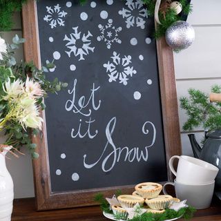 Blackboard with Let It Snow written in chalk, leaning against a white wall on a wooden surface containing flowers, teapot and cups and mince pies