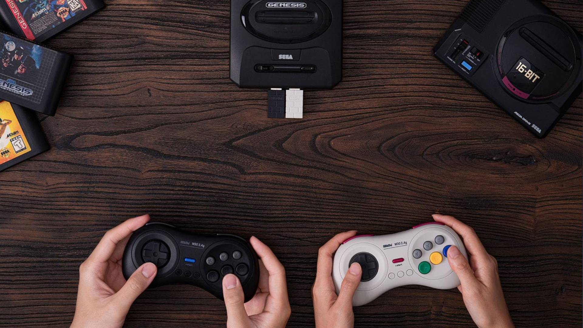 Hands holding 8Bitdo M30 controllers in black and white over woodgrain table next to Genesis Mini.