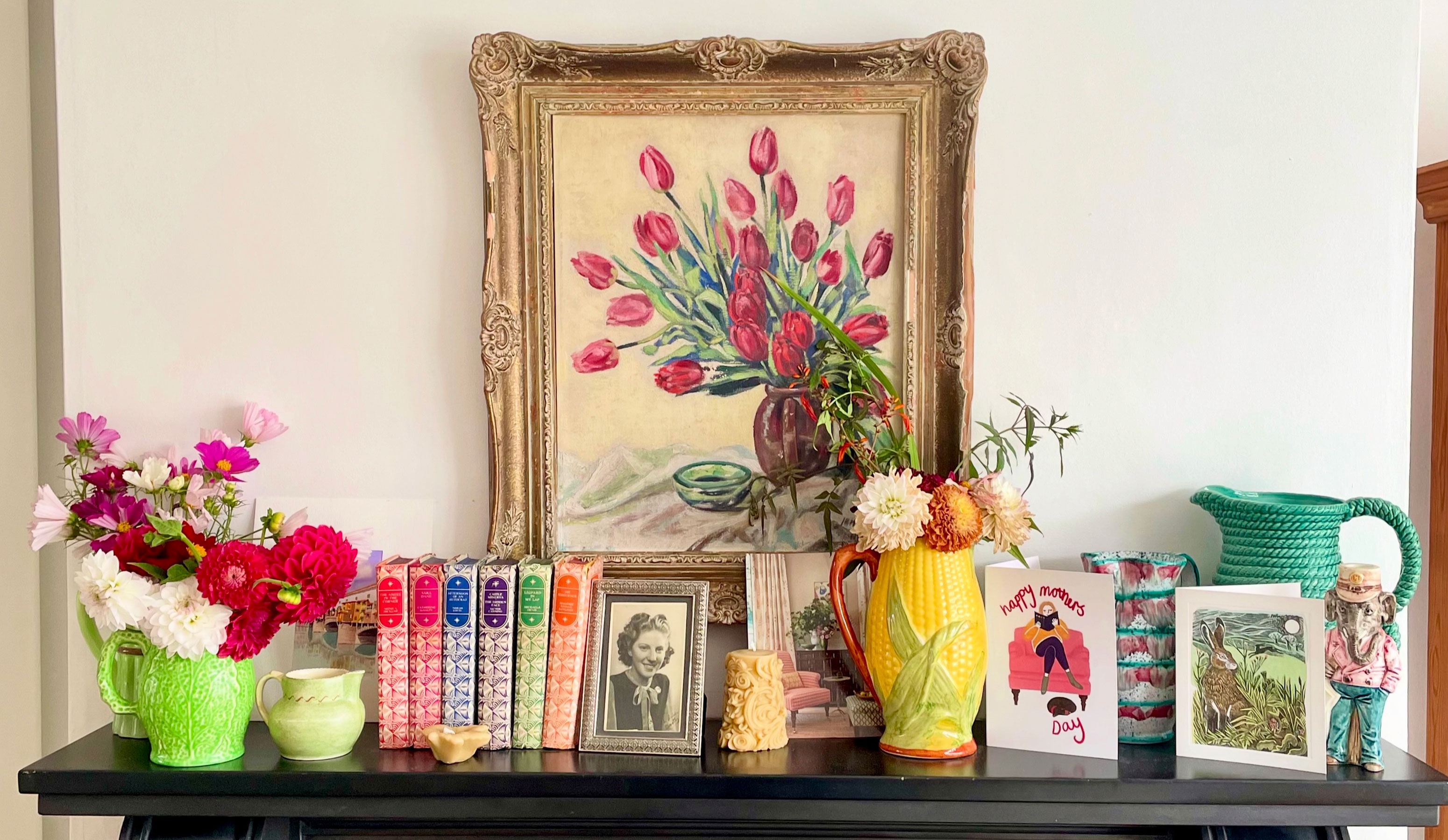 Fireplace mantlepiece with colourful books, vases filled with flowers and floral painting