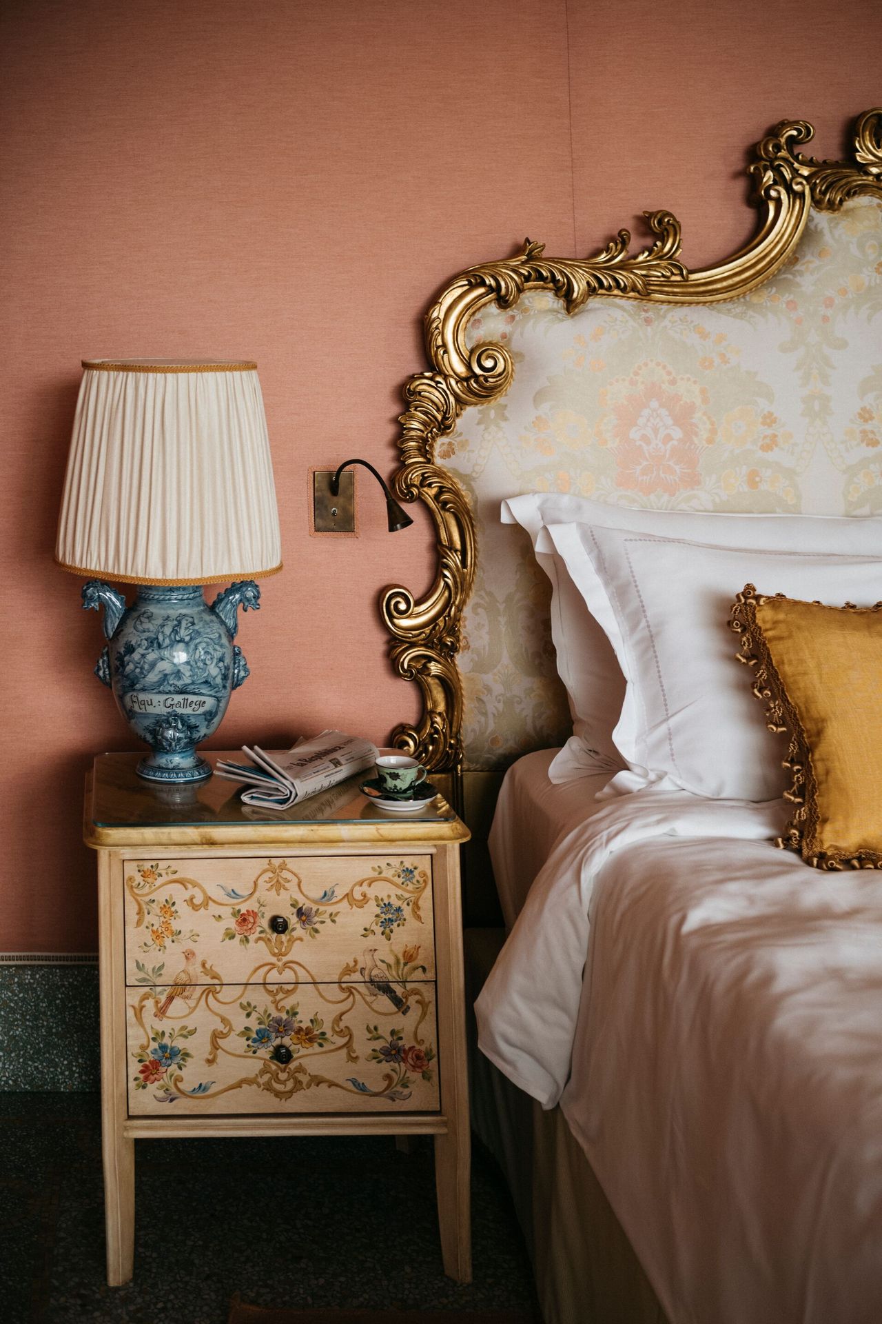 A whimsically painted bedside table with a blue vintage ceramic lamp beside a bed with a gold gilt headboard.