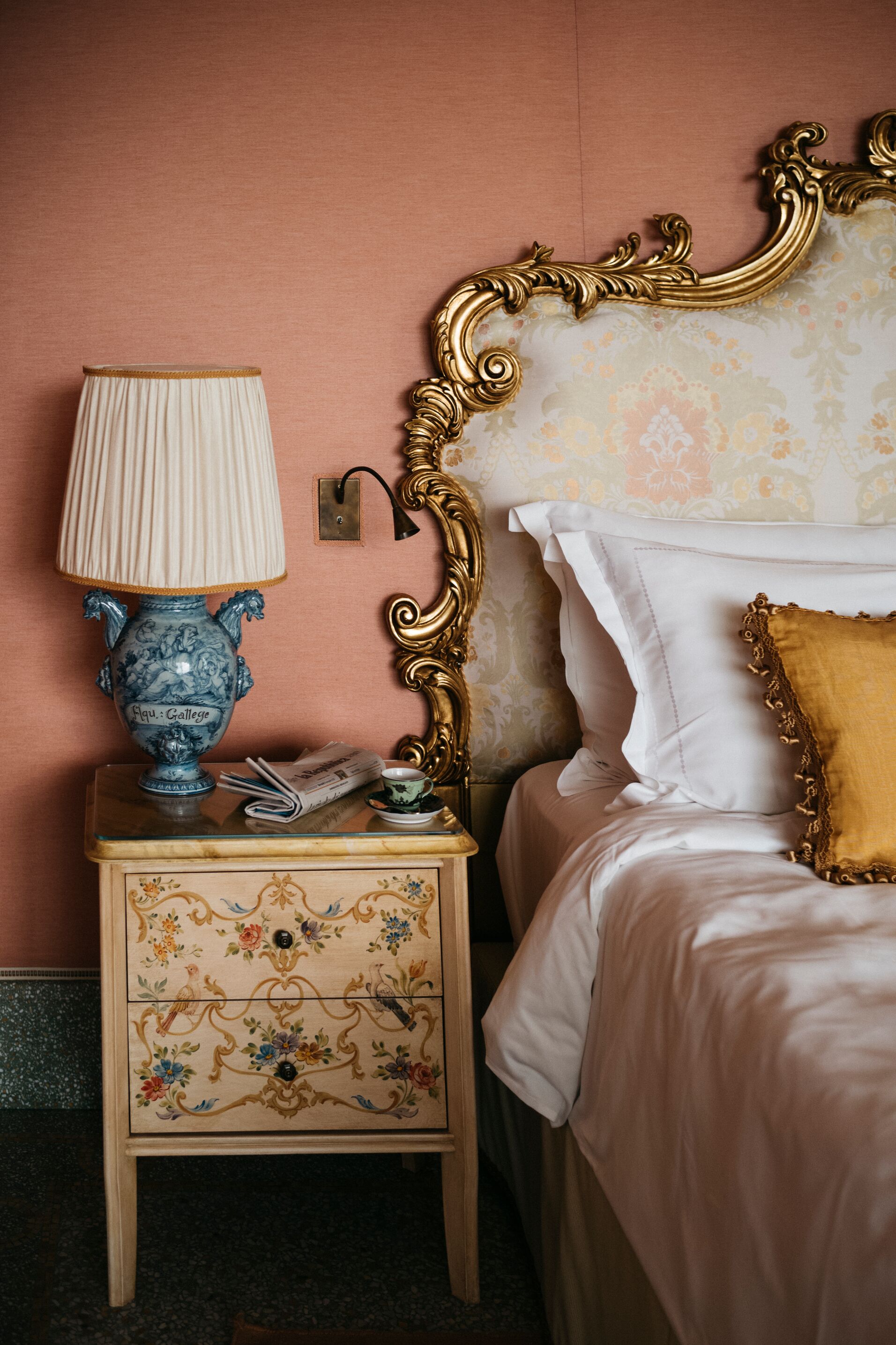 A whimsically painted bedside table with a blue vintage ceramic lamp beside a bed with a gold gilt headboard.