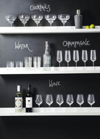 Close up of a range of different drinking glasses, wine bottles and a cocktail shaker on ledges against a black wall.