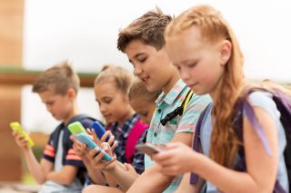 Happy elementary school students with smartphones and backpacks sitting on bench outdoors texting. 