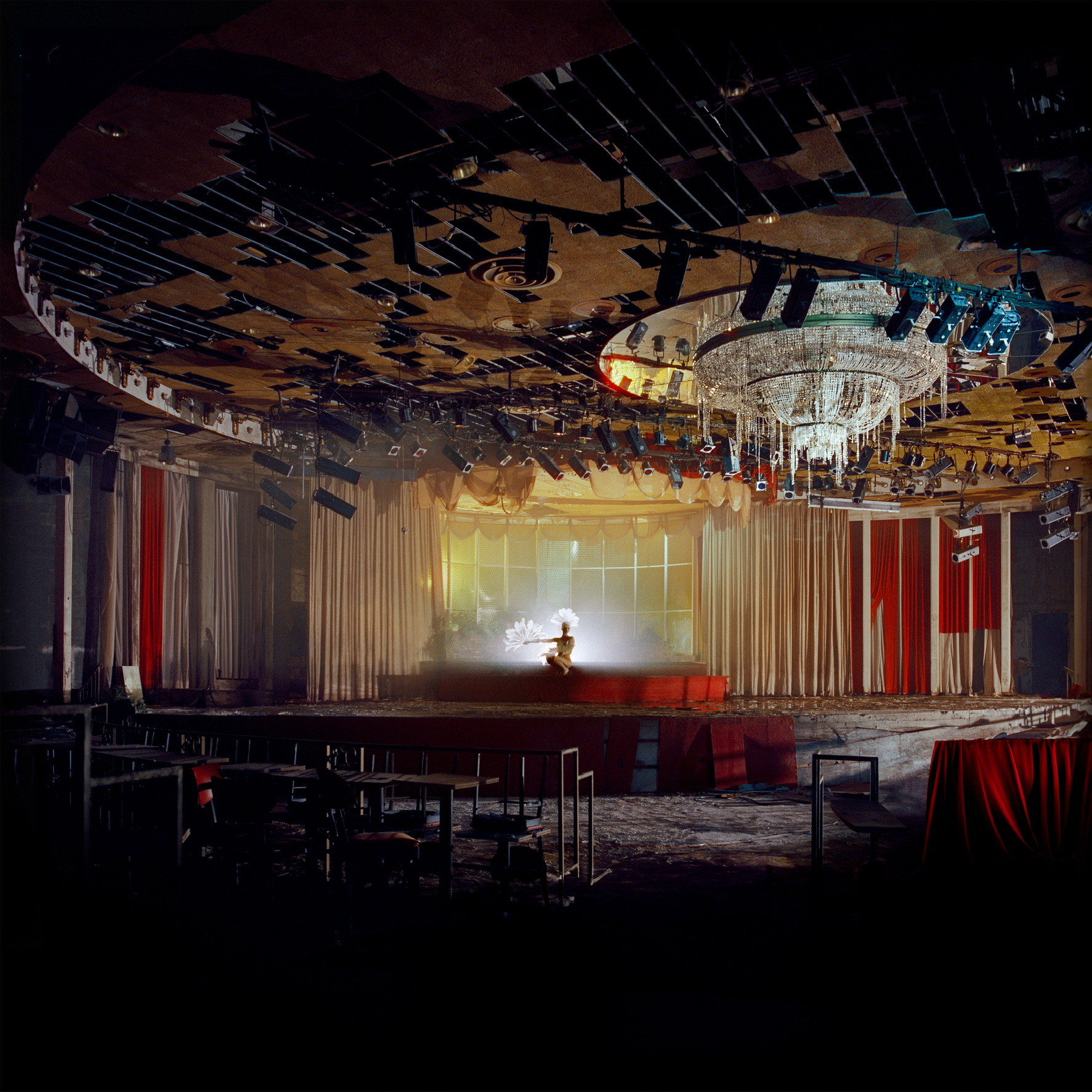 An abandoned theater in a mid-century hotel features a semi-destroyed ceiling, burning red textiles, and a wide chandelier.