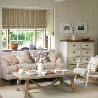 Neutral living room with a wooden coffee table and cream and red striped sofa with Roman blinds hung in the windows and a chest of drawers and armchair