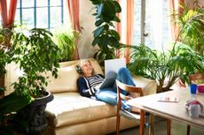 A woman relaxes on a couch surrounded by plants with a laptop.
