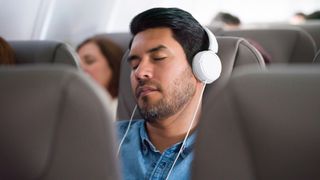 A man with short dark hair wears white headphones to block out noise while he sleeps on a busy plane