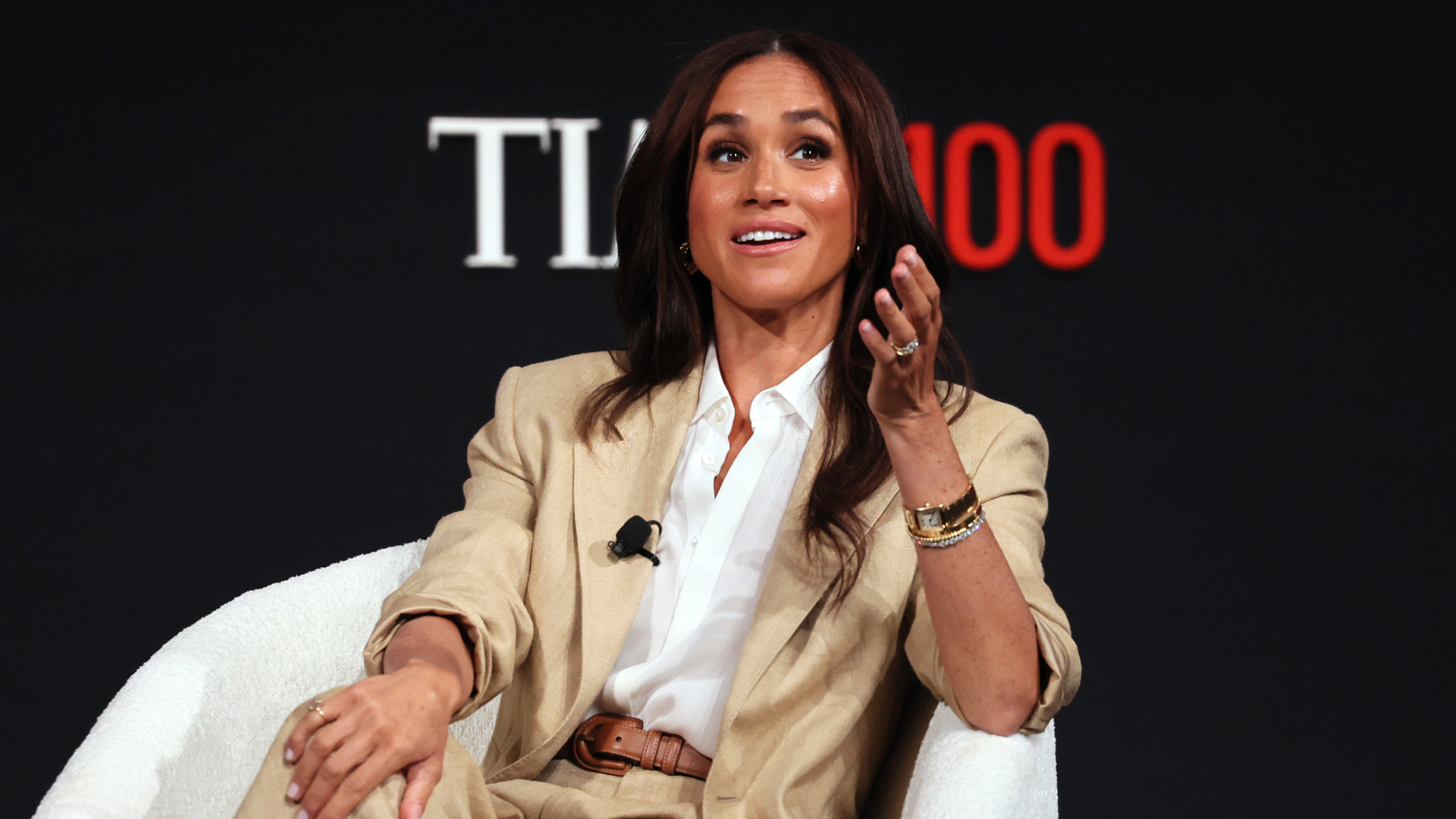 Meghan Markle wearing a tan suit sitting in a chair talking