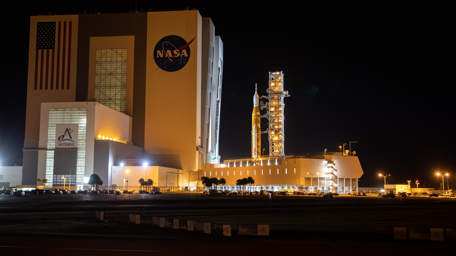 NASA's Artemis 1 Space Launch System moon rocket emerging from the massive Vehicle Assembly Building at night under bright lights.