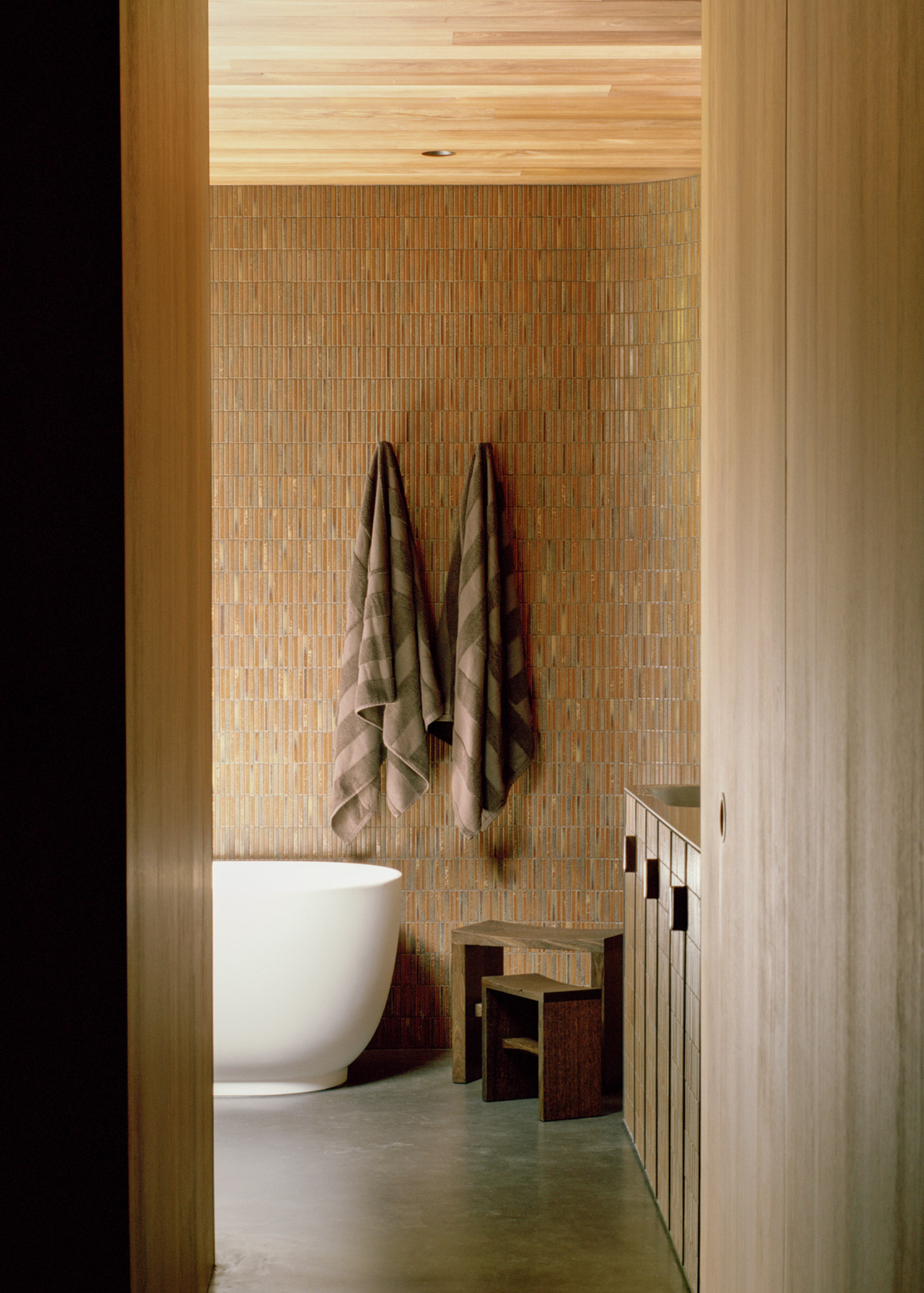 A bathroom with a tiled wall, hanging striped towels, wood stools, and a bath tub