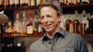Seth Meyers stands behind a bar in a grey shirt while day drinking with Paul Rudd. The Brim of Rudd's cowboy hat is in the corner of the frame.