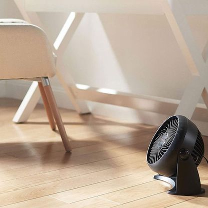 A compact black fan sat on a wooden floor by the side of a white desk and chair